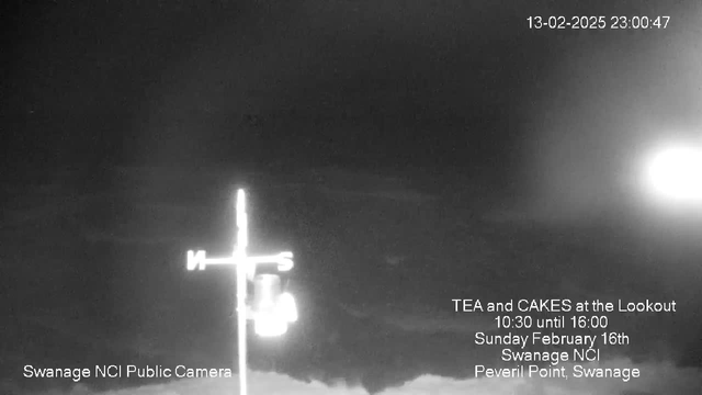 A dark image showcasing a cloudy sky with a small, illuminated area on the right side. In the foreground, there's a weather vane with cardinal direction markers (N, S, E, W) at the top. Text is visible on the bottom left corner indicating "Swanage NCI Public Camera." Additional text in the center provides event details: "TEA and CAKES at the Lookout 10:30 until 16:00 Sunday February 16th Swanage NCI Peveril Point, Swanage." The overall tone is monochrome with limited contrast.