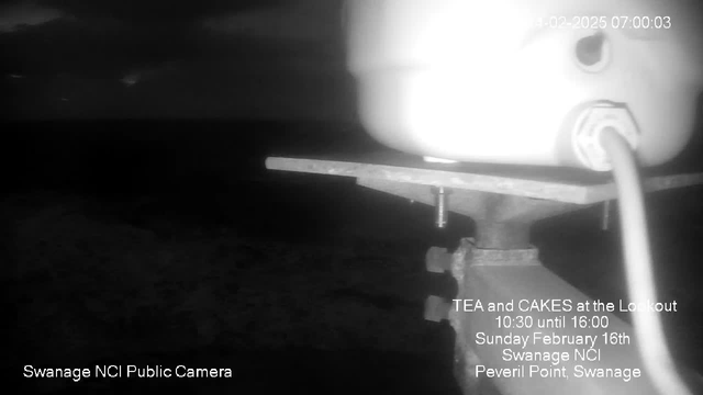 A black and white webcam image showing a metal railing or platform in the foreground with a white object, possibly a canister or container, on top. The background features a dark, cloudy sky with no visible landmarks. Text in the lower left corner indicates the camera is labeled "Swanage NCI Public Camera," while the central area has event information about "TEA and CAKES at the Lookout," including the date and time. The overall scene is dimly lit, creating a moody atmosphere.