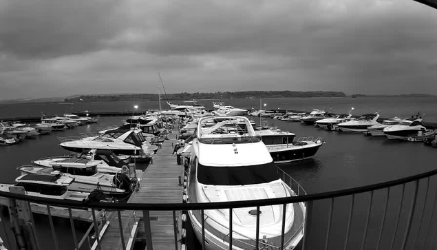 A marina scene with numerous boats docked along a wooden pier. The sky is overcast and gray, reflecting a moody atmosphere. In the foreground, a large white motorboat is prominent, while several smaller boats are visible in the background. The water appears calm, and there are faint outlines of land in the distance. The overall image is in black and white, with varying shades of gray.