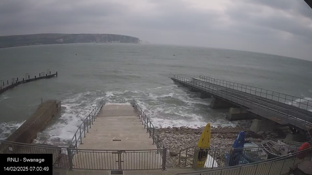 A cloudy scene depicting a shoreline with turbulent waves lapping against a rocky beach. Steps lead down from a metal railing to the water. To the right, there is a wooden pier extending into the sea. Kayaks in yellow and blue are stored nearby on the ground. The distant cliffs are visible in the background, shrouded in mist.