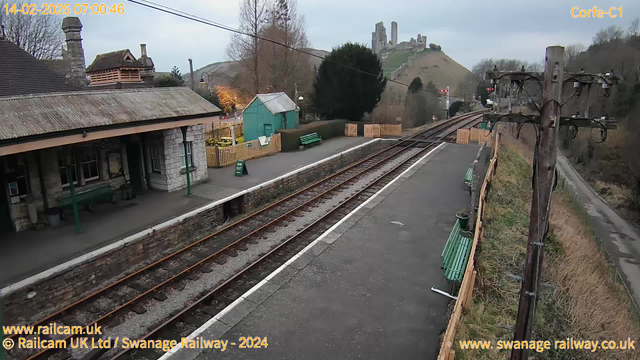 A view of a train station with two railway tracks running parallel. On the left, there is a stone building with a sloping roof and a green bench along the platform. To the right, another green bench is visible, alongside a wooden fence. In the background, a hill rises with the remains of a castle-like structure perched on top. The sky is cloudy, and the scene conveys a calm, early morning atmosphere.