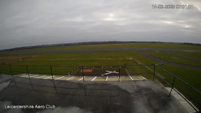 A view from a webcam at the Leicestershire Aero Club, showing a wide expanse of grass-covered land with a runway in the background. The sky is overcast with grey clouds. In the foreground, there's a railing and part of the building rooftop. A red flag is visible further down on the left side, indicating a possible wind direction. The time displayed reads 07:01 on February 14, 2025.