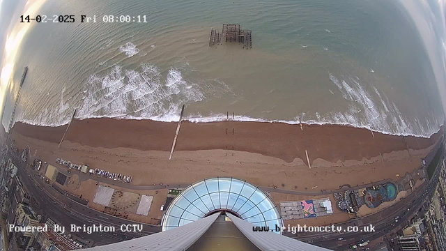 A bird's-eye view of a beach with a sandy shore and rolling waves. The ocean appears in shades of blue and green, with foamy whitecaps along the shoreline. In the distance, a partially submerged pier is visible. Along the beach, there are various structures, including an amusement area with colorful designs, and parked boats. The image has a timestamp indicating the date and time at the top, and part of the photo shows the base of a tall structure with a glass dome.