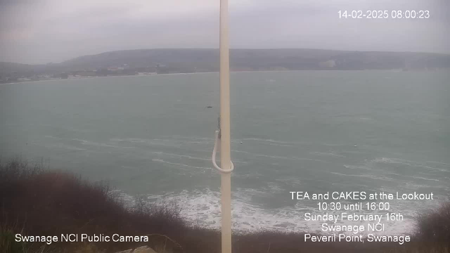 A view from a high lookout point over a choppy sea, with gray skies creating a dull atmosphere. In the foreground, there is a tall, white pole, and to the right, text announces an event for tea and cakes at a lookout. The ocean is rough, with waves breaking against the shore, and the land in the distance features a faint outline of hills mixed with buildings. The scene conveys a sense of a cold and windy day by the coast.