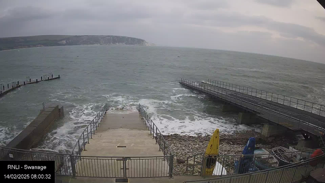 A coastal scene shows turbulent gray waters meeting a rocky shoreline under a cloudy sky. In the foreground, there is a concrete path leading down to the water, flanked by metal railings. To the right, there are two boats on the shore, one yellow and one blue, alongside a pile of various other boats. In the distance, a pier extends into the sea, with additional railings visible. The atmosphere appears overcast and breezy.