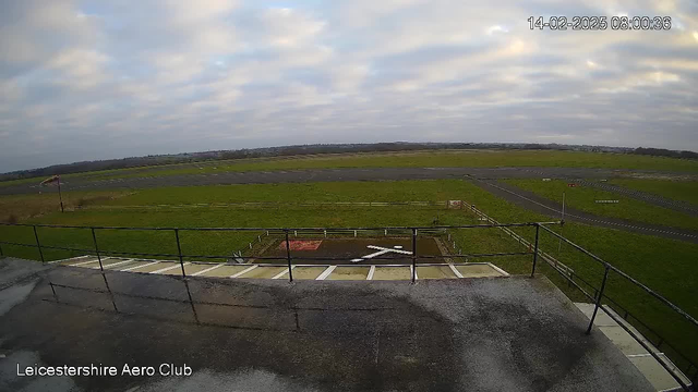 A view from the Leicestershire Aero Club webcam showing a grassy airstrip with a small white "X" marking on the ground. The scene has a gray, overcast sky with scattered clouds. In the distance, there are more grassy areas and a few dirt paths. A flagpole is visible on the left side, and a fence surrounds the airstrip area. The timestamp in the corner indicates the image was taken on February 14, 2025, at 8:00 AM.