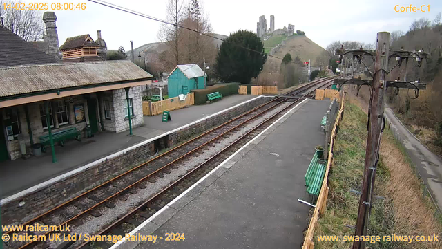 The image shows a railway station platform with green benches and a stone building on one side. There are train tracks stretching into the distance on the left, with a hilly landscape behind them. A wooden fence separates the platform from an adjacent area. In the background, there is a hill topped with castle ruins. The sky appears overcast with gray clouds, and there are trees in the vicinity. A wooden pole with electrical wires is visible on the right side of the image.