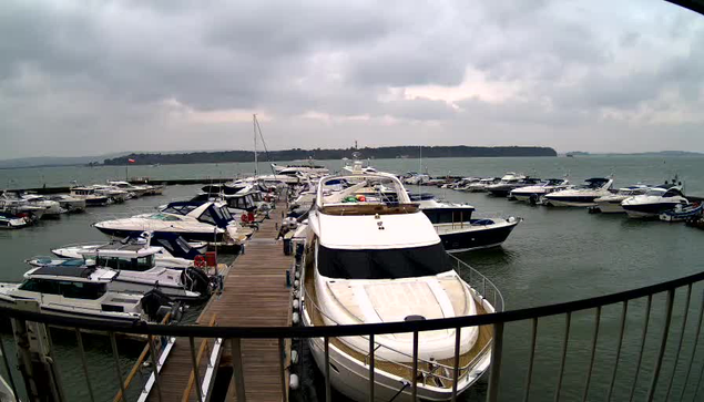 A view of a marina on a cloudy day. Numerous boats and yachts are docked along a wooden pier. The water appears calm with slight ripples, and a few boats are visible in the distance. The sky is overcast, contributing to a muted atmosphere.