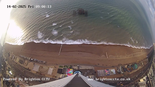 Aerial view of a sandy beach with gentle waves lapping at the shore. To the left, an old pier is partially submerged in the water, and several boats are visible nearby. The beach features colorful structures and attractions, along with a promenade and pathway leading along the coast. The scene is bright with morning light reflecting on the water, creating a shimmering effect. The image also includes date and time stamps at the top right corner.