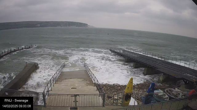 A cloudy, overcast scene depicts choppy waters along a coastline. In the foreground, a set of stairs leads down to the sea, flanked by railings. To the right, there is a wooden pier extending into the water. The waves crash against the shore and pier, creating white froth. On the side, a few kayaks are visible on the rocky beach, with one yellow kayak positioned upright. In the background, cliffs rise in the distance under the gray sky.