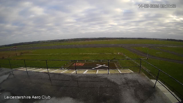 A view from a high vantage point overlooking an airfield. The foreground features a flat rooftop with a railing, while an expansive green field stretches out in the middle ground. There are markings on the ground, including a large white "X" and a small circular area. In the distance, there are more sections of tarmac and a few trees nearby, with a cloudy sky overhead. The scene appears tranquil and open, characteristic of an aerodrome environment.