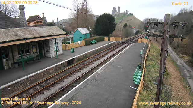 A railway station scene with tracks running horizontally across the image. On the left, there is a stone train station building with a gray roof, green benches, and a poster on the wall. A green building is visible behind the station. To the right, the railway tracks lead into the distance, bordered by a wooden fence. In the background, there are trees and a hill with ruins at the top, suggesting a castle or fort. The sky is gray, indicating an overcast day. Additional benches are placed along the platform, and a sign saying "WAY OUT" is positioned on the ground. A power pole with various wires stands to the right of the image.