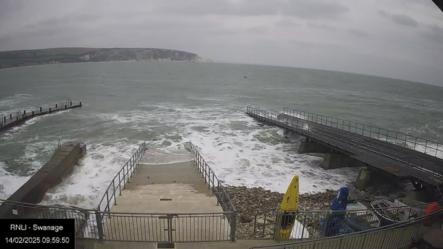 A coastal scene showing turbulent waves crashing against a rocky shore. In the foreground, there are steps leading down to the water, flanked by railings. To the right, a small wooden jetty extends out over the water, while on the left, a section of the shore appears concrete. Two kayaks, one yellow and one blue, are positioned on the beach. The sky is cloudy, and a distant cliff can be seen in the background, partially obscured by the clouds.