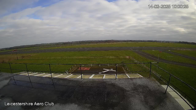 A view from a webcam showing a grassy airfield with a runway. There is a large "X" marking on the ground at the center of the image, surrounded by white boundary posts. The sky is partly cloudy, and a distant horizon is visible. The date and time are displayed in the top right corner, indicating February 14, 2025, at 10:00:25. A railing is visible in the foreground.