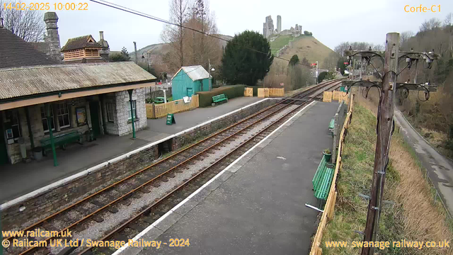 A view of a railway station with a platform on the left. The station building, made of stone with a corrugated roof, is visible with green benches nearby. In the background, a hilly landscape features a historic castle ruin atop a hill. On the right, railway tracks stretch into the distance, lined with grass and shrubs. A wooden utility pole with wires stands beside the tracks, and a sign indicates "WAY OUT" near a picket fence. The sky is overcast, creating a muted atmosphere.