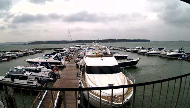 A view of a marina on a cloudy day. Various boats and yachts are docked in the water, with a wooden walkway extending along the edge. The water appears slightly choppy, and the horizon includes a distant landmass. The scene conveys a calm marine atmosphere but is overshadowed by the overcast sky.