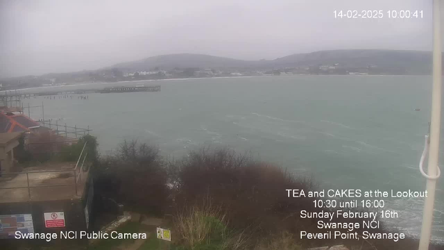 A cloudy view of a coastal landscape featuring a calm sea with small waves. In the foreground, there are some bushes and a sign with caution messages. A wooden pier extends into the water, and behind it, a sandy beach is visible. In the distance, there are buildings and hills shrouded in mist. The date and time are displayed in the corner, indicating it is 10:00 AM on February 14th, 2025. Text on the image mentions a tea and cakes event at Swanage NCI.