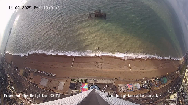 Aerial view of a beach scene with light brown sand, gentle ocean waves, and a cloudy sky. In the foreground, the image shows the tip of a tall structure. Below, there are people walking on the beach, along with some beachside attractions and structures, including a circular area with colorful designs and a pool. An old pier can be seen in the water, partially submerged. The image also displays timestamps at the top left corner and a watermark at the bottom.