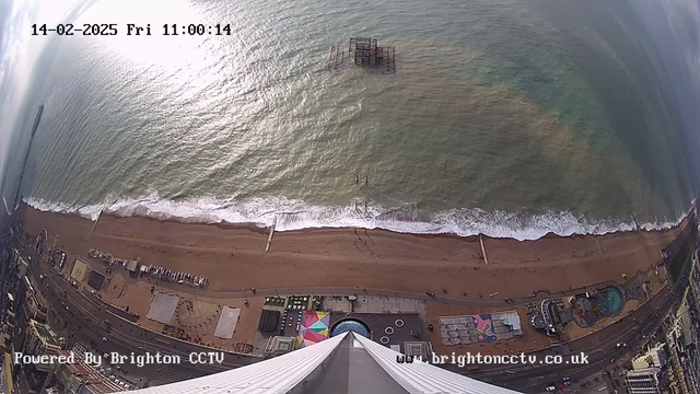 An aerial view of a beach during daylight, showing a sandy shore and waves breaking at the water's edge. In the water, a partially submerged structure is visible, possibly remnants of a pier. The beach is lined with a promenade, and several small structures, including rides and cafes, can be seen near the waterfront. The sky is clear with some clouds, and sunlight reflects off the water. In the lower section, the image features a sharp drop from the viewpoint, with the edge of the viewing structure visible, leading down to the beach. The time stamp in the top left indicates February 14, 2025, at 11:00:14.