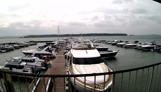 A view of a marina filled with various boats and yachts. The scene captures a wooden dock extending into the water, lined with several white and blue vessels. The water is calm, and the sky is overcast, creating a grayish atmosphere. In the background, more boats are visible, and a distant shoreline can be seen, suggesting a natural landscape beyond the marina.