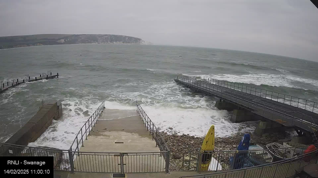 A view of a coastal area with choppy gray waters. There is a rocky shore in the foreground leading to a set of stairs that descend toward the water. To the right, there are two colorful kayaks, one yellow and one blue, resting near the water's edge. A wooden pier extends into the water in the background, partially covered by waves. The sky is overcast, and there are distant cliffs visible along the shoreline.