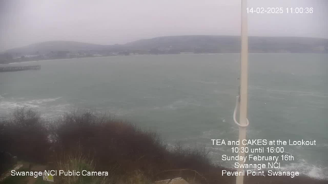 A cloudy, overcast view of the sea at Peveril Point, Swanage. The ocean appears choppy with whitecaps, and a wooden pier is visible on the left side of the image. The shoreline is lined with dark green foliage and sparse bushes in the foreground. In the bottom left corner, there is text indicating it is a public camera for Swanage NCI, while the right side has information about a tea and cakes event happening on February 16th. The date and time in the top corner notes February 14, 2025, at 11:00:36.