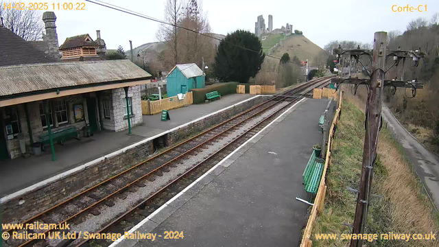 A view of a railway station platform taken from a webcam. The foreground features a wide, empty platform with a few green benches lining the side. In the background, there is a low stone wall bordering the railway tracks, which are visible running parallel to the platform. A wooden telegraph pole with wires stands to the right of the image. In the distance, a hill rises up, topped by castle ruins. The sky appears cloudy, giving the scene a muted light.