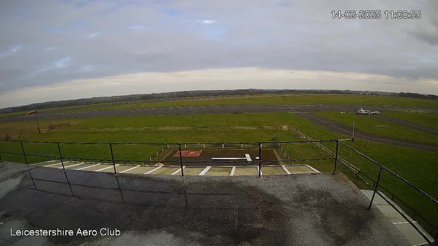 A view from a webcam shows a grassy airfield under a cloudy sky. In the foreground, there is a railing and a wet surface, likely from rain. In the middle ground, an airplane is parked on the tarmac, and part of the runway is visible. The area is bordered by a wooden fence, and the landscape is open with some distant trees along the horizon. The scene captures a calm atmosphere typical of an airport setting.