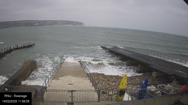 A seaside view showing turbulent gray waves crashing against a rocky shoreline. A set of stairs leads down to the water, flanked by a railing. To the right, there are two brightly colored kayaks: one yellow and one blue, resting on a sandy area. A wooden pier extends into the water nearby, with a second, smaller section visible further out. The sky is overcast, and the landscape in the background features steep cliffs.