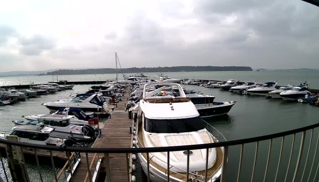 A view of a marina filled with various boats and yachts docked at a wooden pier. The scene is set under a cloudy sky, with some boats displaying colorful canopies. In the background, a calm body of water reflects the gray clouds, and a distant shoreline is visible. The area has a peaceful atmosphere, and the arrangement of the boats suggests a busy yet orderly marina.