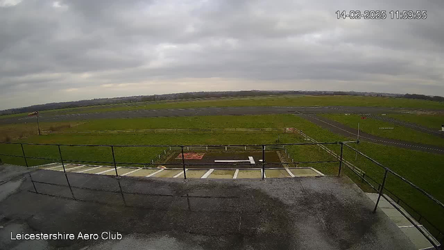 A wide view of the Leicestershire Aero Club's airfield, featuring a grassy area with a few scattered trees in the background. The sky is overcast with gray clouds. In the foreground, a railing borders a small observation deck. The runway stretches out, displaying marked lanes and a paved taxiway. A windsock on a pole indicates wind direction, and there are distant hills visible on the horizon.