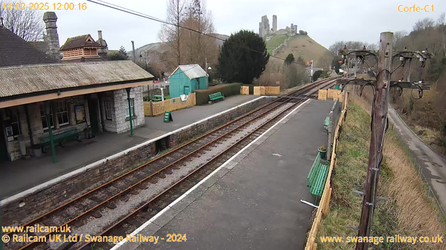 A view of a train station with a stone platform. There are several green benches along the platform. On one side, a small wooden shed is visible, painted light blue. The station building has a sloping roof and a stone wall. In the background, a green hill rises, topped with castle ruins. The sky is cloudy, and a power pole with wires stands to the right, partially obscured by trees. The railway tracks run parallel to the platform, leading into the distance.