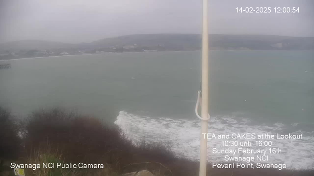 A cloudy and overcast view of the sea from a high vantage point, showing choppy greenish waters with white waves breaking at the shore. In the foreground, there are dark bushes and grasses, while the blurred outline of a coastal town and hills can be seen in the distance. A white flagpole stands on the right side of the image, with soft white clouds in the gray sky. Text at the bottom provides information about a local event.