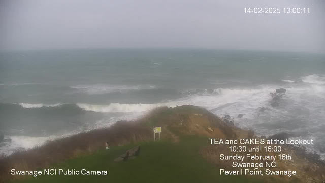 A seaside view from a webcam showing turbulent waves crashing against a rocky shore. The sky is overcast, and the water appears choppy, with shades of blue and gray. In the foreground, there is a grassy area with some benches and a sign. The image includes text advertising a tea and cake event at the Lookout on February 16th, from 10:30 to 16:00, along with a timestamp indicating the date and time of the image.