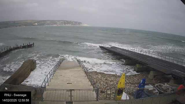 A view of a coastal area showing a set of concrete steps leading down to the water. Waves are crashing against the rocky shoreline, and a small pier extends into the sea on the right side of the image. Two colorful kayaks, one yellow and one blue, are positioned at the edge of the pier. The sky is overcast with clouds, and the surrounding hills are visible in the background. The scene gives a sense of a windy day by the seaside.