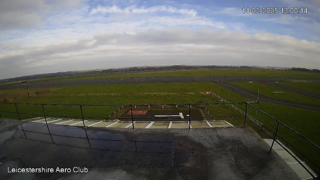 A view from a webcam at Leicestershire Aero Club shows a vast, grassy landscape under an overcast sky. In the foreground, there is a railing on a deck, with steps leading down. Below, a tarmac runway is visible, with a small section of white markings indicating a landing strip. The area surrounding the runway is fenced, with patches of grass and some signs. The sky is mostly cloudy, creating a grayish atmosphere. The scene appears calm with no visible aircraft.