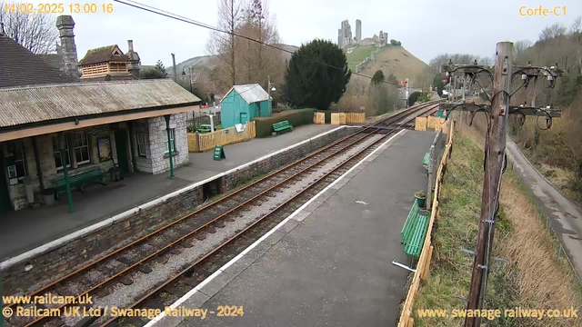 A railway station scene featuring a partially covered platform with stone walls and a canopy overhead. Green benches are positioned along the platform. To the left, there is a small green shed and a section of the station is visible, including a sign that reads "Way Out." In the background, a steep hill is seen with a castle ruins on top. The railway tracks run alongside the platform, leading into the distance. A wooden utility pole is on the right, with wires and equipment attached. The sky is overcast.