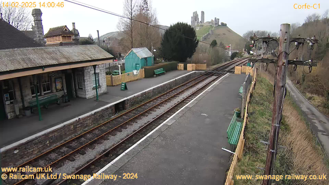 A railway station scene with a stone building featuring a sloped roof and wooden benches along the platform. In the background, a hill can be seen topped with ruins of a castle. There are green benches and a wooden fence surrounding the area, along with a small blue shed and traffic signals. It's a cloudy day with a clear view of the tracks and surrounding landscape.