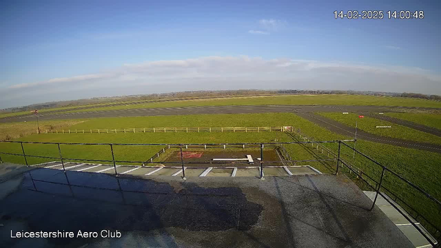 A wide view from a webcam at Leicestershire Aero Club shows a grassy landscape under a clear blue sky. In the foreground, a railing is visible, suggesting a viewing platform. Below, there is a rectangular area with a wet surface, possibly a puddle, surrounded by a white fence. The background features a flat, green field with a runway marked by a dark asphalt surface and a few signs indicating directions. The timestamp in the corner reads 14:00:48 on February 14, 2025.