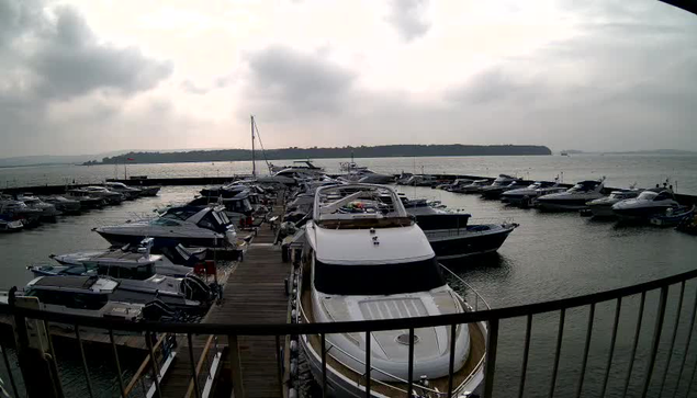 A marina scene featuring numerous boats docked at a wooden pier. Several yachts and smaller boats are visible, with some parked closely together. The water is calm, reflecting the cloudy sky above. In the background, a hillside is partially obscured by the clouds, and there appears to be a faint outline of land on the horizon. The overall atmosphere is tranquil, with a muted color palette of gray and blue.