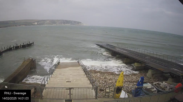 A cloudy day at a coastal area, featuring a stone walkway leading down to the sea, bordered by metal railings. To the left, a wooden pier extends into the water, with several pilings visible. Waves crash against the shore and the walkway, with smooth stones scattered along the beach. On the right, colorful kayaks in yellow and blue are stacked near a small dock. In the background, a cliff can be seen along the coastline. The time displayed is 14:01:03 on February 14, 2025.