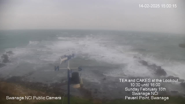 A cloudy, gray scene of the ocean on a windy day. Waves crash against dark rocks at the shoreline, creating white froth. In the foreground, there is a weather vane pointing south and a sign indicating the location of Swanage NCI Public Camera. The date and time are displayed in the top right corner, and text about a tea and cakes event is visible on the right side of the image. The atmosphere appears stormy and moody.