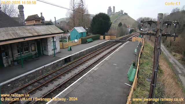 A railway station scene with a rustic stone building on the left featuring a sloped, tiled roof. Green benches line the platform, and there are wooden fences separating the area. In the background, there is a small blue shed with a sloped roof and trees nearby. Train tracks stretch along the platform, leading toward a distant hill that hosts ruins or a castle. The sky is overcast, and electrical poles are along the side of the tracks.