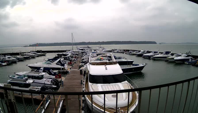 A marina scene on an overcast day, featuring numerous boats docked in the water. A wooden dock leads into the marina, surrounded by various motorboats and yachts, some with blue and white color schemes. The sky is gray with clouds, and the water appears calm, extending towards the horizon where land is faintly visible.