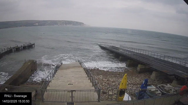 A coastal scene featuring a rocky shoreline with gentle waves lapping at the edge. There are two piers extending into the water; one is a solid structure, while the other has a wooden walkway. Stairs lead down to the water from a concrete area with a railing. To the right, several colorful kayaks are stored near the water, with orange, yellow, and blue visible. The sky is overcast, and a distant coastline is faintly visible in the background.