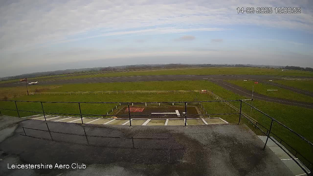 A view from a webcam overlooking a runway at Leicestershire Aero Club. In the foreground, there is a railing and part of a structure, while the runway extends into a grassy area. There are several markings and a few parked small aircraft visible on the runway. The sky is mostly cloudy with a hint of blue. The surrounding landscape is open, with fields in the distance.