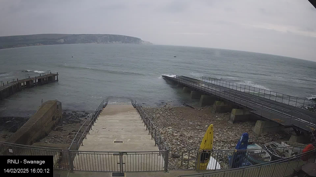 A view of a shoreline with water lapping at the edge. There is a set of stairs leading down to the beach, bordered by a metal railing. To the left, a small jetty extends into the water, and in the foreground, there are a few brightly colored kayaks. The landscape includes rocky textures and a backdrop of cliffs in the distance under a cloudy sky. The scene conveys a calm coastal atmosphere.