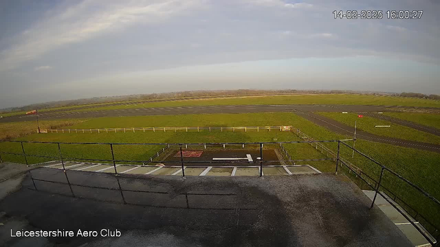 A wide-angle view from the rooftop of Leicestershire Aero Club, showing a grass airfield with a gray runway marked by white lines. In the foreground, there is a railing surrounding a flat rooftop area. A windsock stands to the left, indicating wind direction. The background features a cloudy sky and distant hills, while the airfield is bordered by a white fence. The image appears to be taken in daylight.