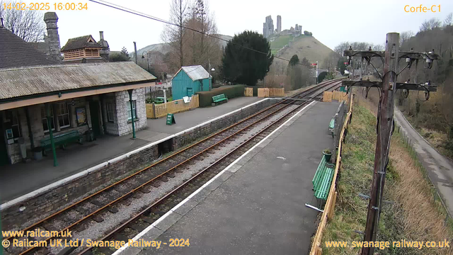 A railway station scene featuring a stone building with a sloped roof and green benches. The platform is lined with tracks leading away into the distance. In the background, there is a hill with a castle ruin. A green shed and several trees are visible near the station. The sky is overcast, and there is a wooden fence separating the platform from the adjacent area. A telephone pole with wires stands on the right side of the image.