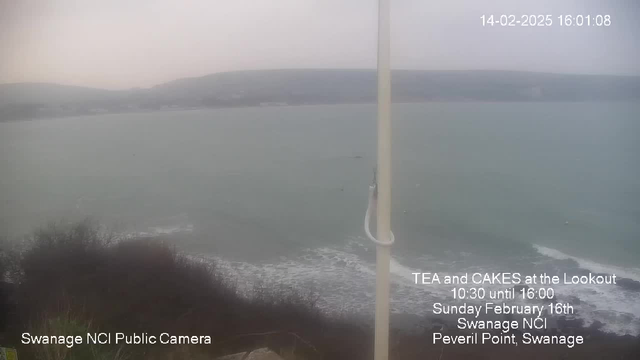 An outdoor seaside view is captured under overcast, gray skies. The scene includes a body of water with gentle waves, a distant shoreline lined with hills. A white pole is on the left side of the image. Text in the foreground advertises a tea and cake event, mentioning specific dates and location details. The overall atmosphere appears calm and subdued, typical of a cloudy day at the coast.